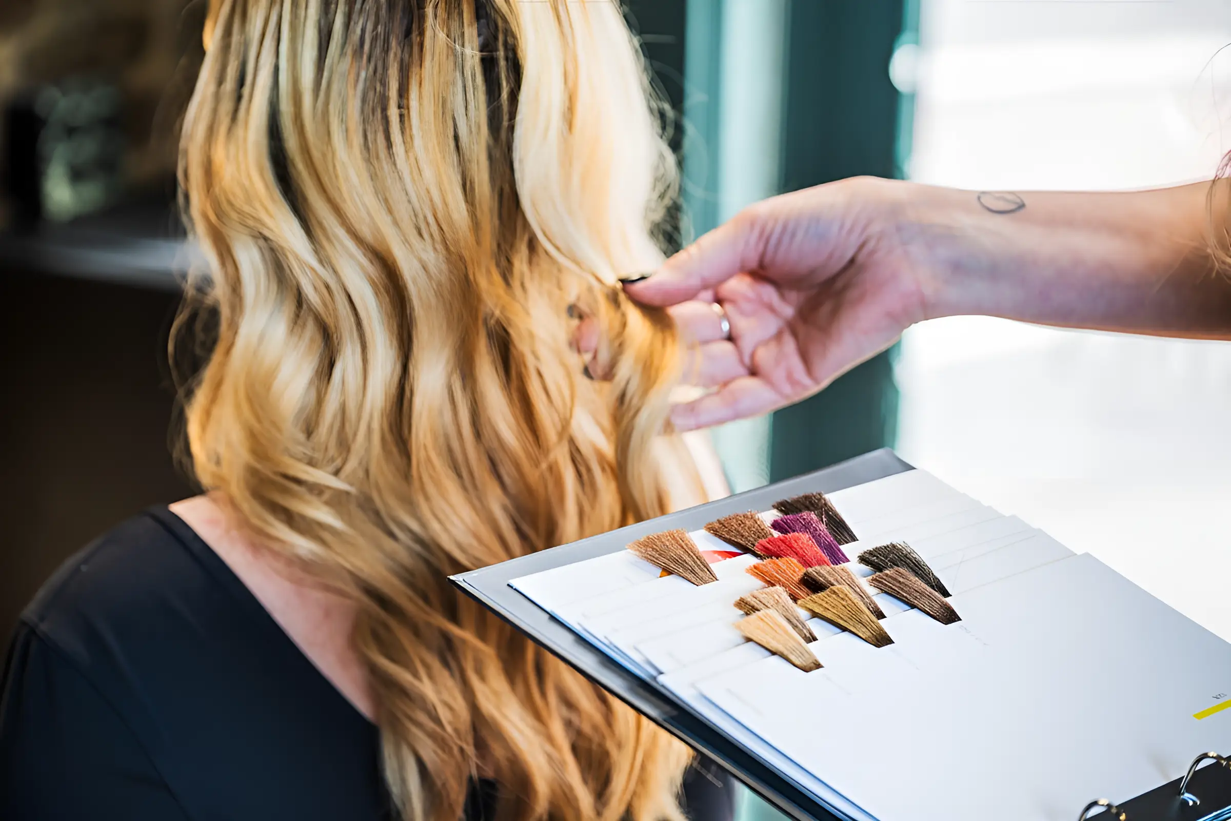 Close-up of a hairstylist’s hand holding a lock of blonde hair against a color swatch book, Lox Salon hair color consultation